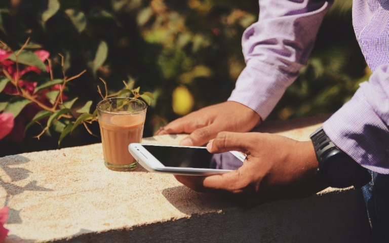 A person sits on a wall in front of plants and looks at their cell phone. 