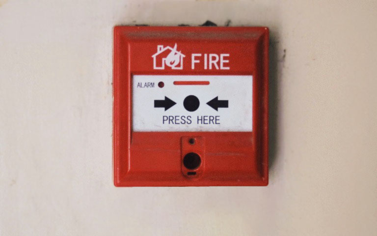 An old, red fire alarm on an off-white building wall