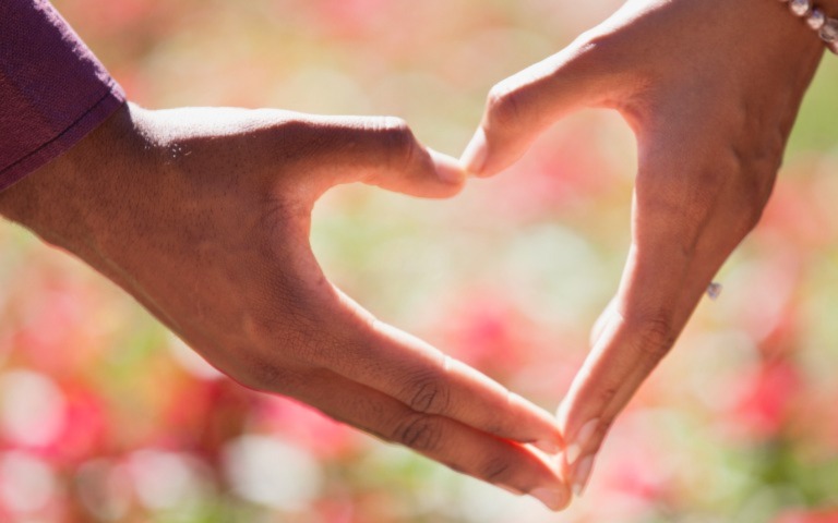 A close up of two hands forming a heart together. Background is sun and grass.
