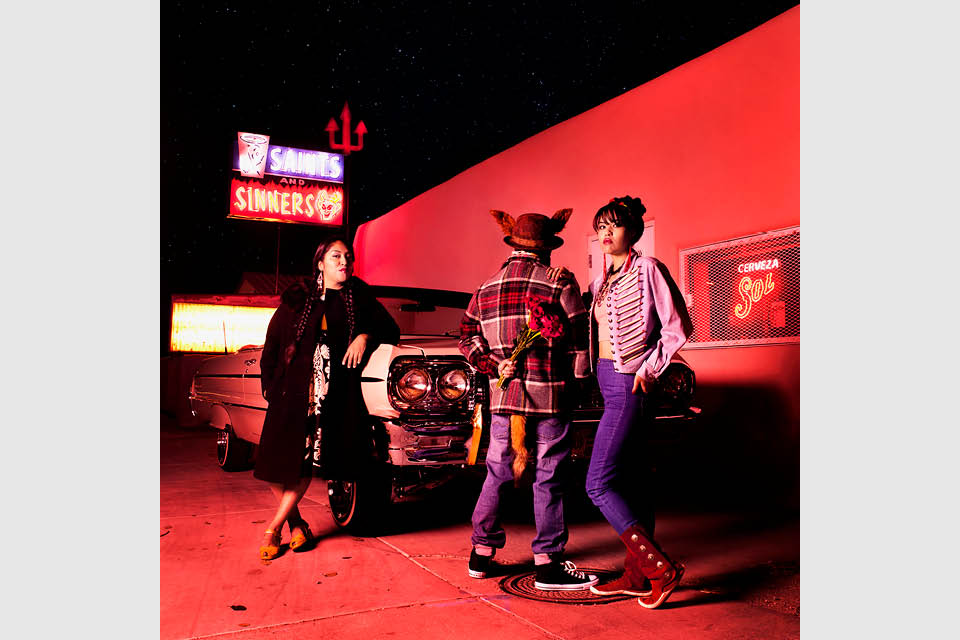 Three women stand next to a vintage car under a sign that says "Saints and Sinners" bathed in a bright red light.