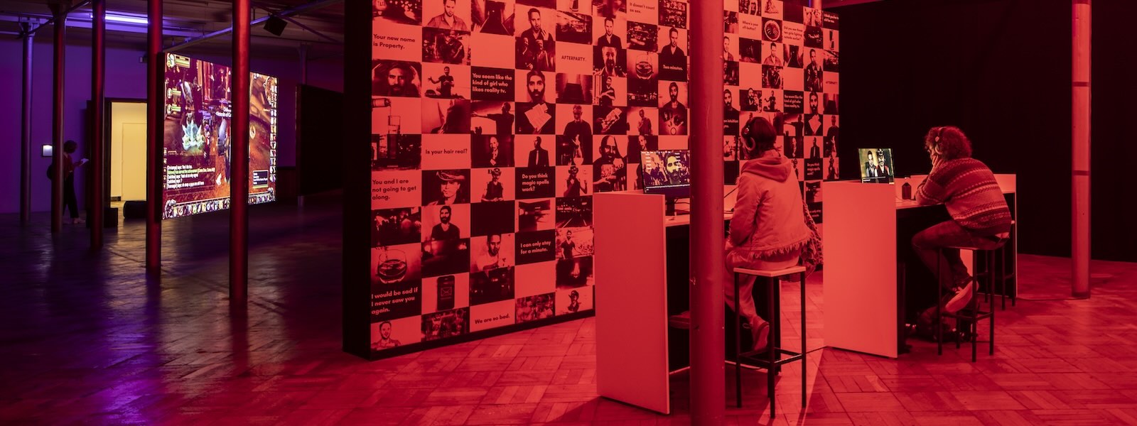 A room bathed in red light with people sitting at computer monitors and facing a wall with various headshots and quotes.
