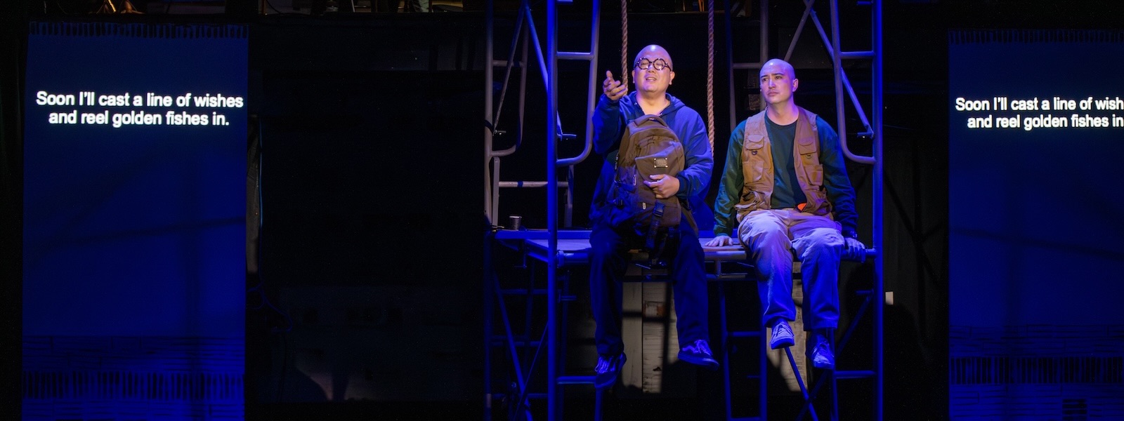 Two men on a theater stage sit on scaffolding. Projected text reads: Soon I'll cast a line of wishes and reel golden fishes in.