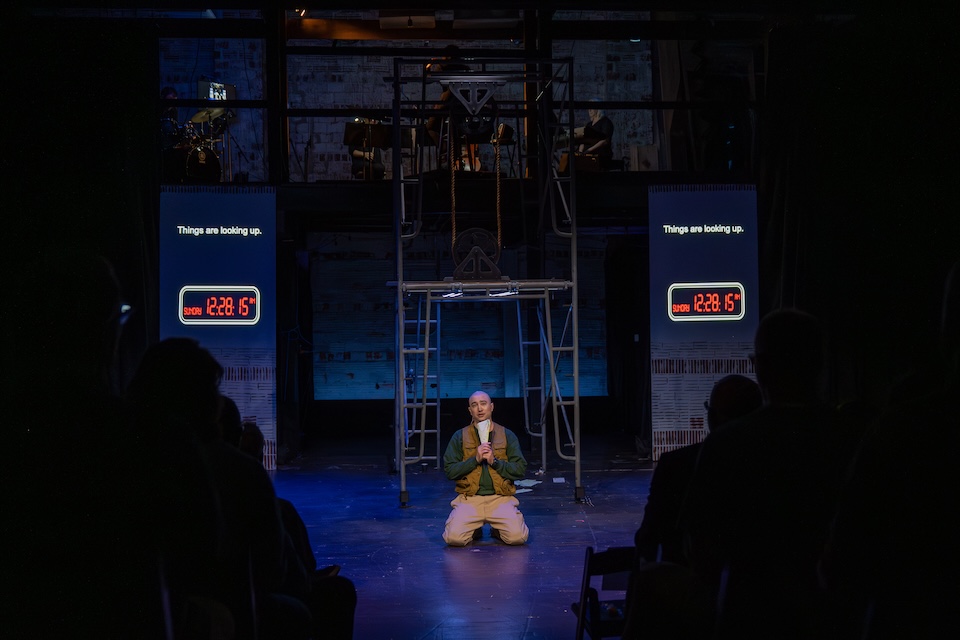 A man on stage is kneeling in front of scaffolding with his hands clasped. Projected text reads: Things are looking up. 12:28:15 PM
