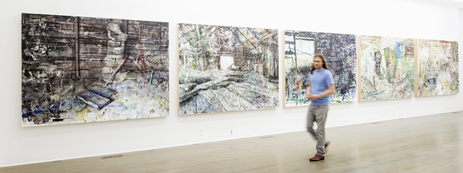 A man walks past a gallery wall with five paintings of abstracted room interiors.