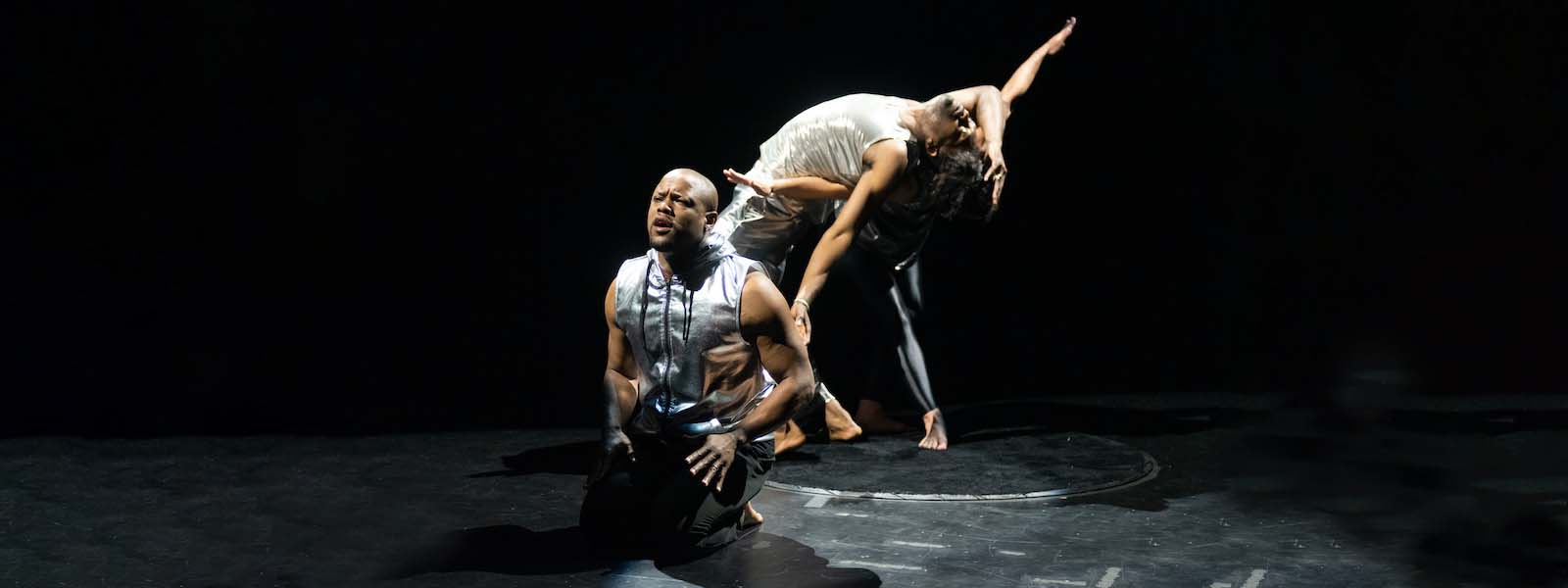 A Black man on stage kneels in front of two dancers in the background.