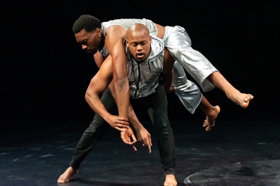 Two dancers are on stage - both Black men - one is carrying the other across his shoulders.