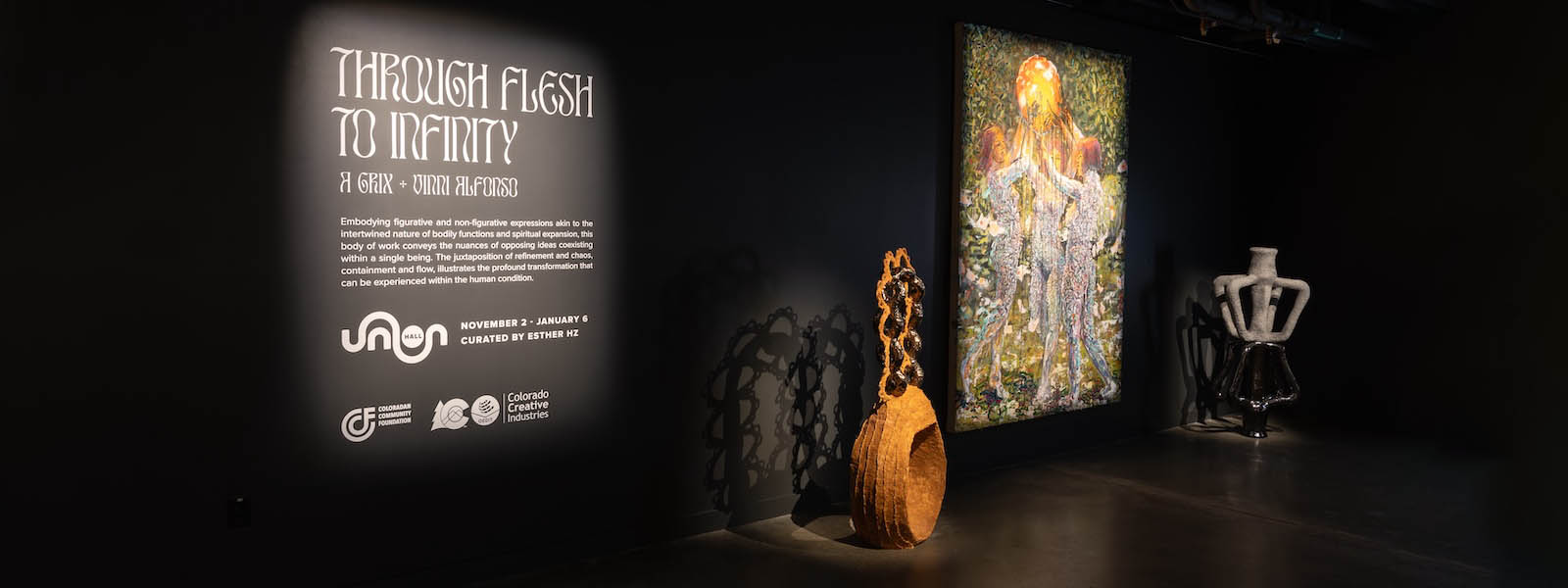 The entrance to an exhibition in a dark room with spotlights on a text panel that reads "Through Flesh to Infinity" and several artworks on the right of it.
