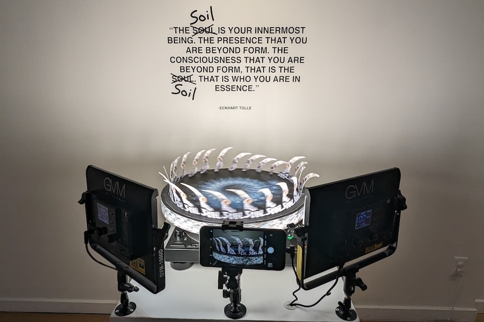 A zoetrope with video screens are in the forground. Text on the wall behind reads witht he word Soul crossed out and Soil written in: " The soul (soil) is your innermost being. The presence that you are beyond form. The consiousness that you are beyond form. That is the soul (soil). That is who you are in essence. - Eckhart Tolle