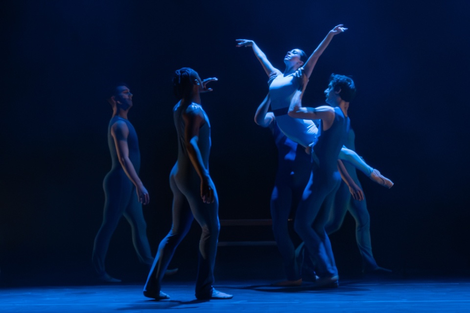 On a dark stage under a blue light, two dancers approach two other dancers who are lifting up a third dancer.