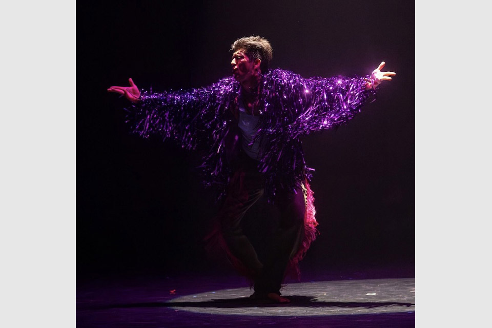 A man onstage with arms outstretched wearing a shimmering purple outift.