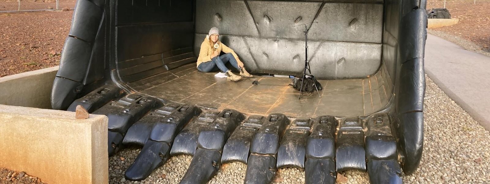 A white woman holding a microphone sits inside a very large excavator bucket scoop.