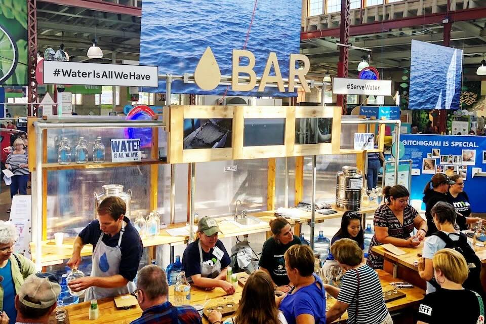 A group of people are being served glasses of water at a temporary bar. A sign reads Water Is All We Have.