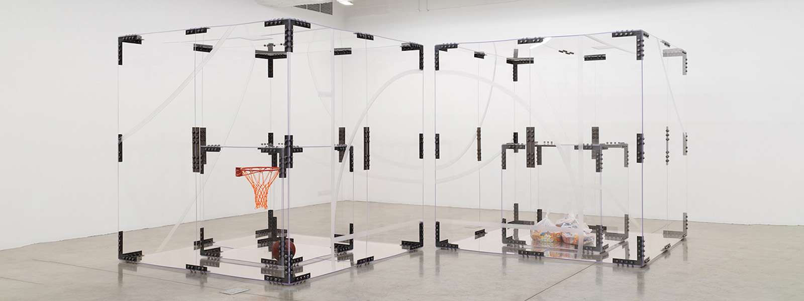 Two large, clear plexiglass boxes in a white gallery space. One box contains a basketball hoop and basketball, the other contains clear plastic bags filled with colorful material.
