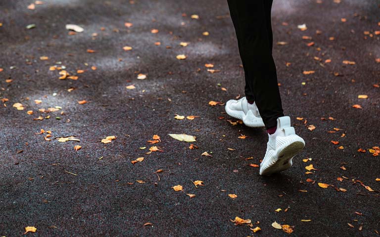 Person in black leggings and white tennis shoes walking on black pavement amongst fall leaves.
