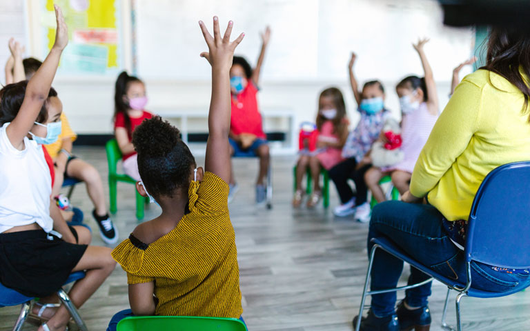 Young children in a classroom sit in a circle raising their hands.