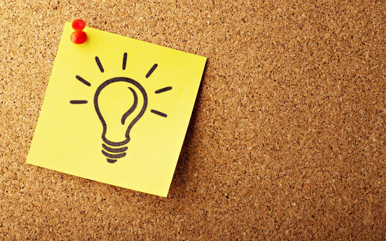 Yellow Post-It note with a lightbulb on it is tacked to a cork board