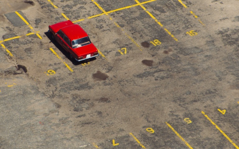 An old, red car sits in an empty parking lot striped in yellow. 