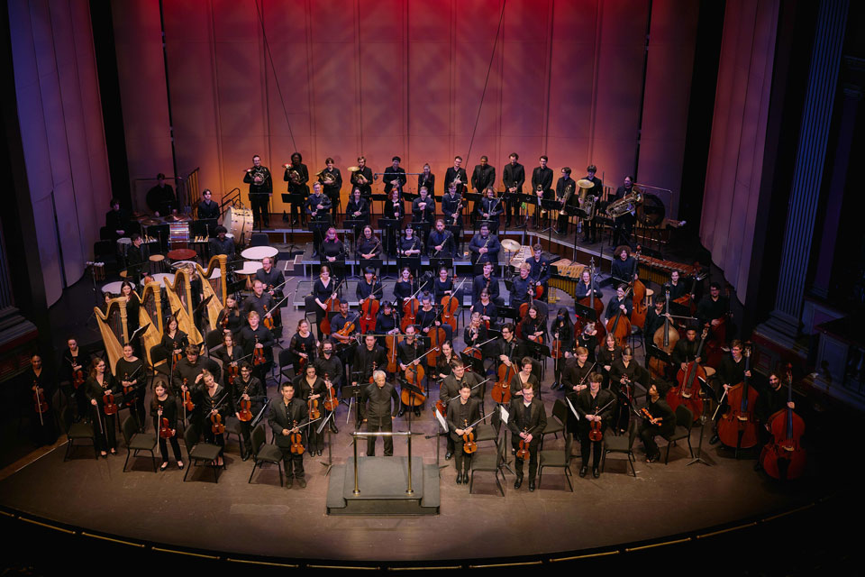 A conductor leads an orchestra in a lively concert performance, with musicians focused on their instruments