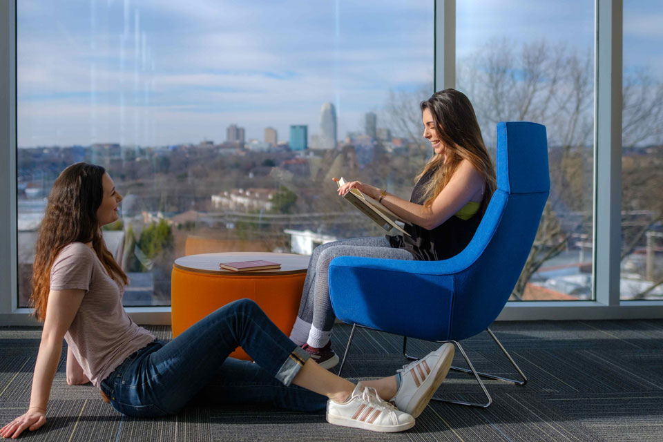 Two women seated in chairs, engaged in conversation, in front of a large window with natural light streaming in.