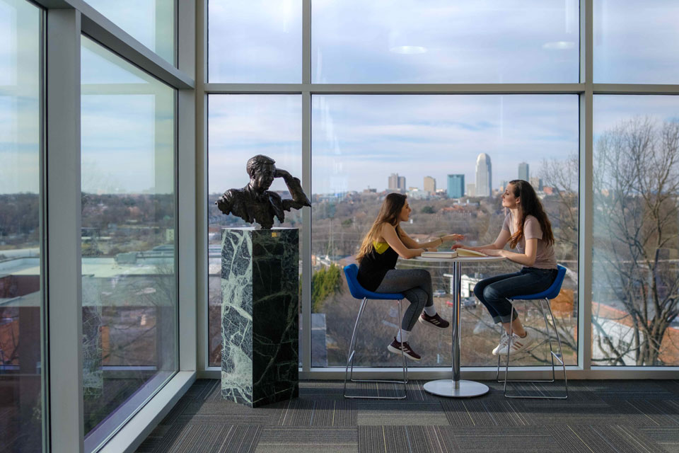 Two women seated at a table, engaged in conversation, with a large window providing natural light behind them.