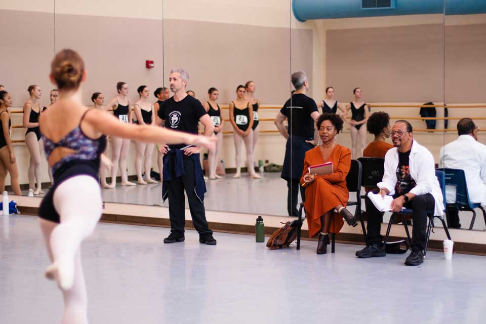 A ballet class in a studio, with students practicing while others observe from the sidelines.