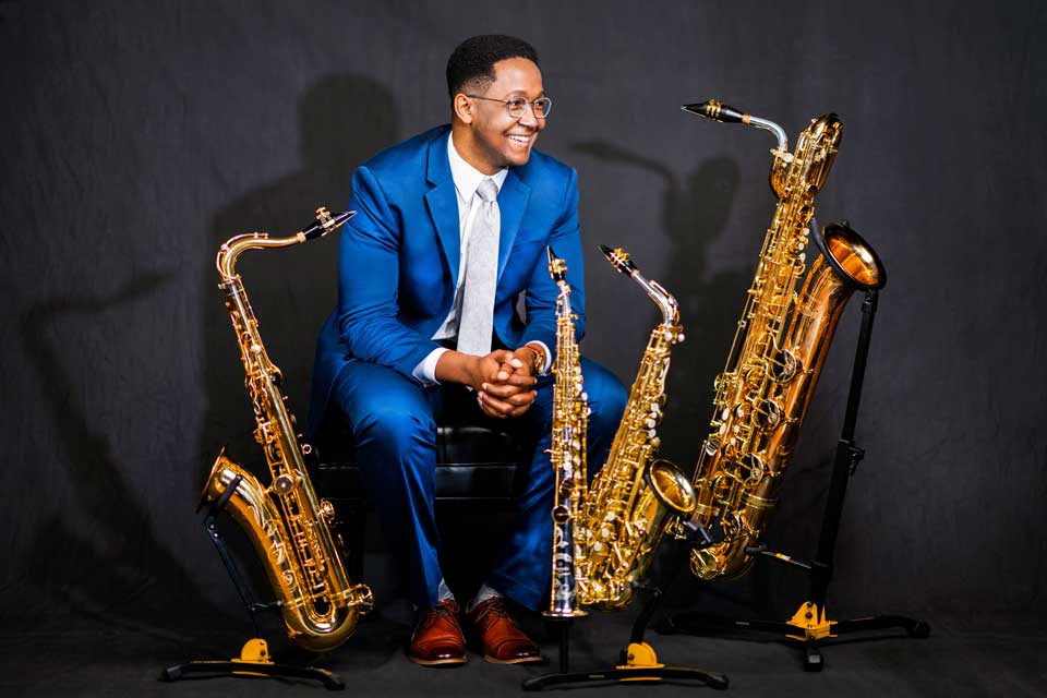 A man in a blue suit and glasses sits beside three saxophones, showcasing a blend of style and musical instruments.