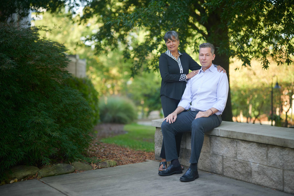 A man and woman sit on a bench, surrounded by greenery, with a large tree in the background.