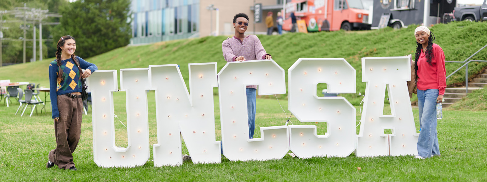 Three smiling students pose outdoors behind UNCSA letters.