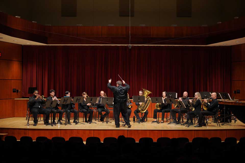 A band performs in a concert hall, led by a conductor, with musicians playing various instruments on stage.