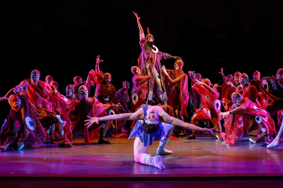 A group of dancers performing energetically on stage, showcasing their choreography and costumes under bright lights.
