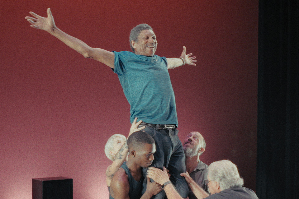 Group lifts smiling dancer on stage against red backdrop