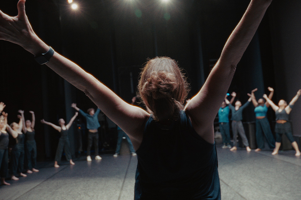 Dancer lifts arms leading group circle on stage under lights