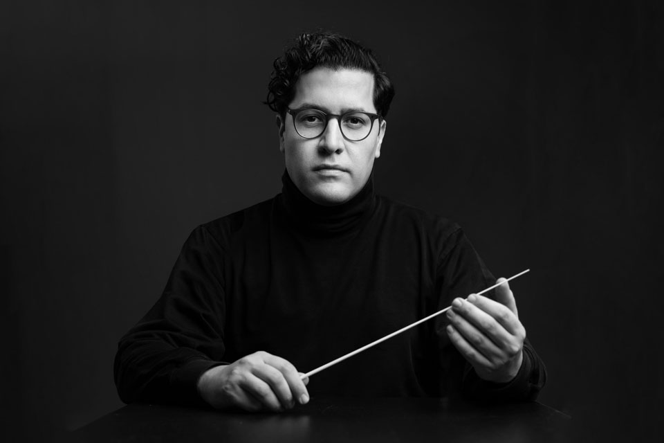 A bespectacled man stands with a stick held out in front, suggesting he is preparing for conducting.