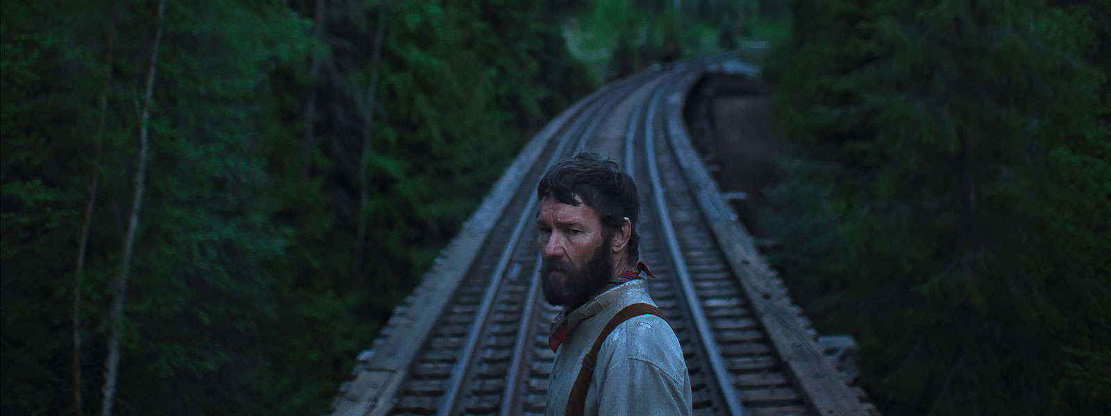 A bearded man stands confidently on a train track, surrounded by greenery and distant train signals.