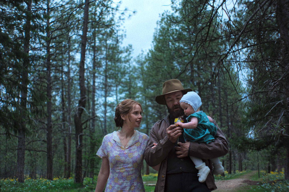 A man and woman hold a baby together, surrounded by lush greenery in a serene forest setting.