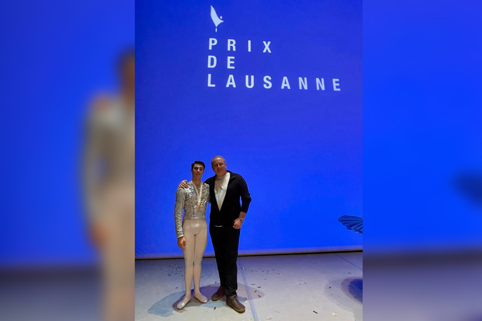 Two people stand beside a blue sign reading "Prix de Lausanne," indicating the location of the prestigious ballet competition.