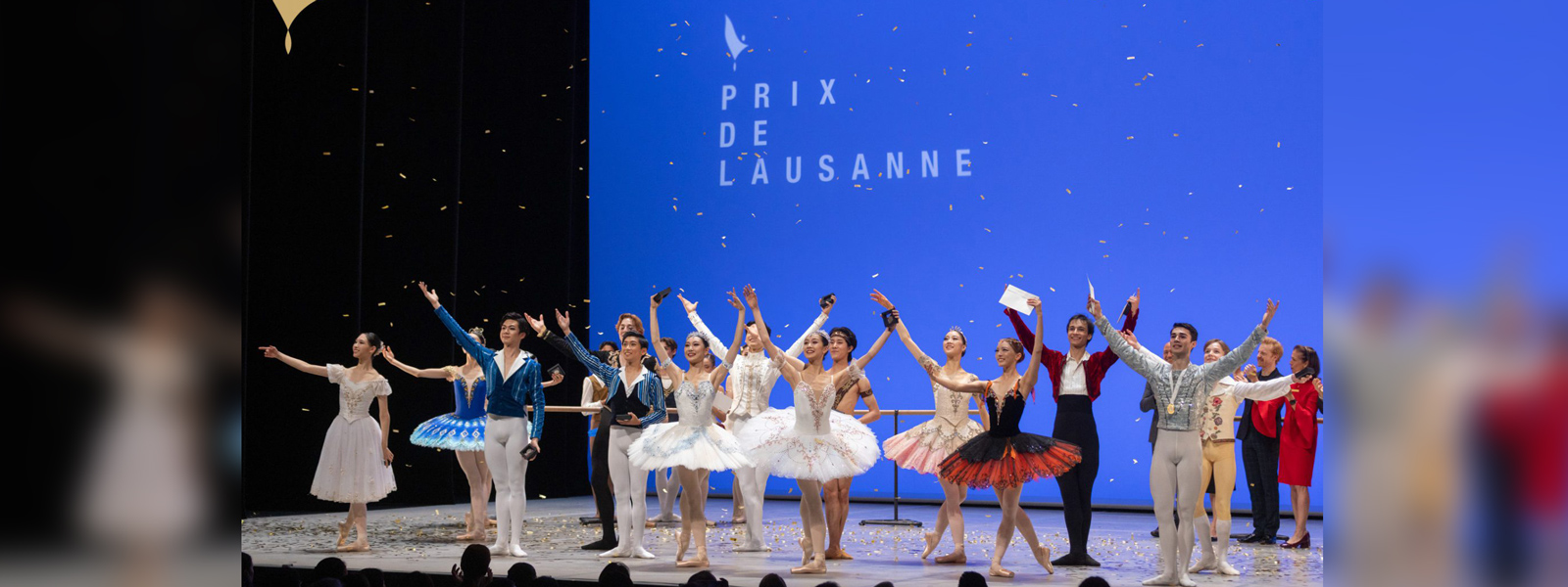 Dancers perform on stage, illuminated by colorful lights, with a large screen displaying visuals behind them.