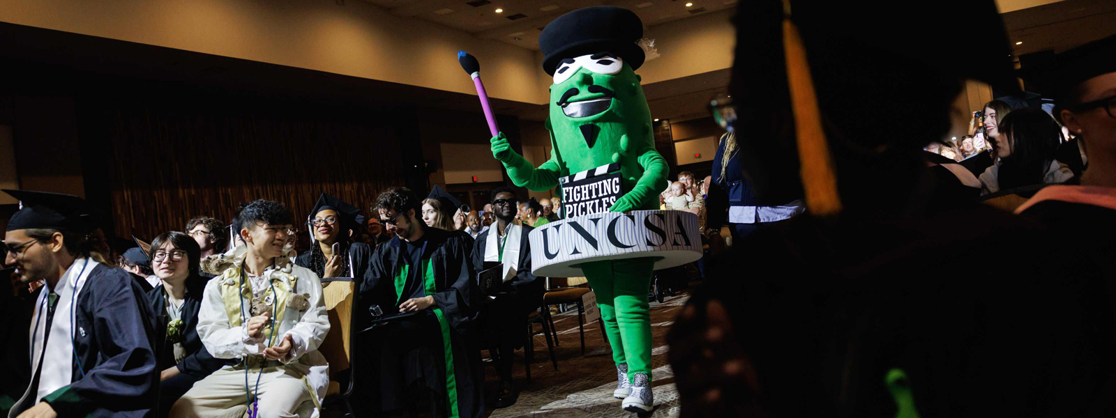 A large green pickle mascot is cheered on by people while wearing a graduation hat