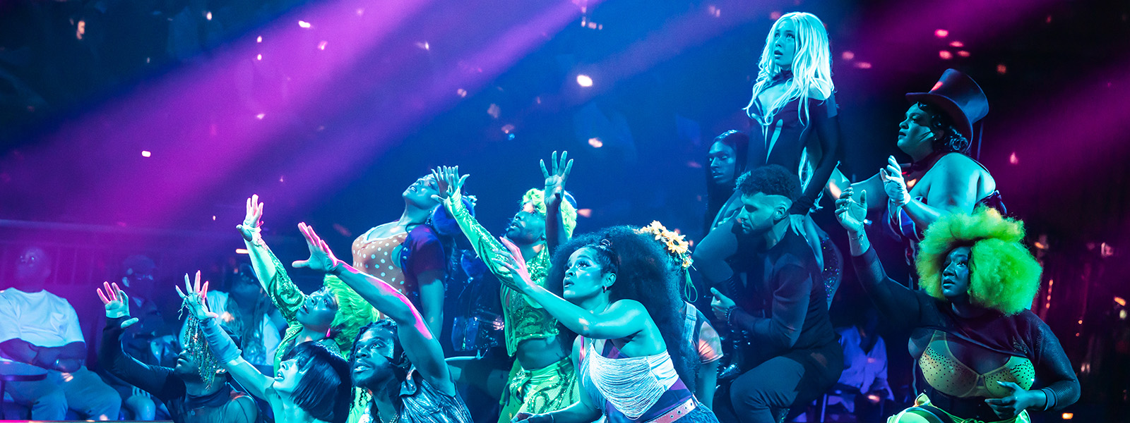 Performers in vibrant costumes reach their arms upward in unison during a high‑energy number from “Cats: The Jellicle Ball.”