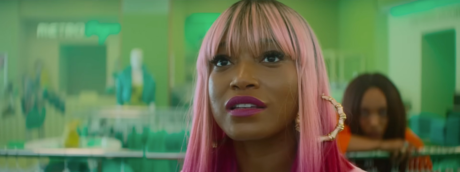 A woman with pink hair standing against a green background with a serious look.